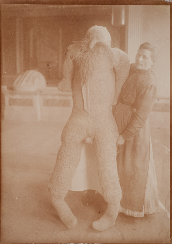 Historische Sepiafotografie: Eine Frau steht neben einer lebensgroßen, aus grobem Textilgewebe gefertigten menschenähnlichen Figur mit ausgestreckten Armen und hält sie im Innenraum eines Gebäudes aufrecht.